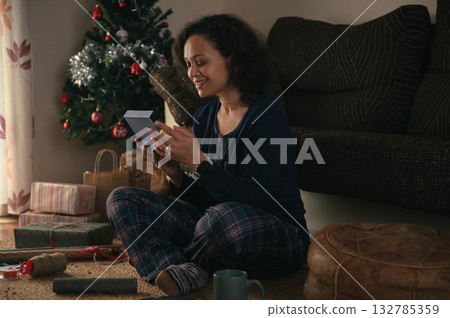 Woman Sits on Floor Unwrapping Gift Near Christmas Tree in Cozy Living Room 132785359