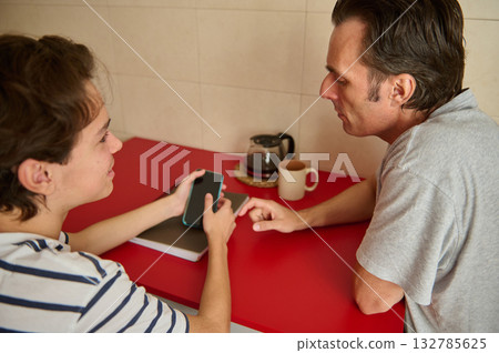 Two Adults Sit Across From Each Other At A Red Kitchen Counter Looking At A Smartphone Two Adults Sit Across From Each Other At A Red Kitchen Counter Looking At A Smartphone 132785625