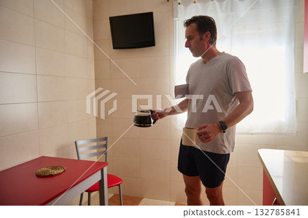 Man In Kitchen Pouring Coffee From Pot While Holding Mug During Morning Routine Man In Kitchen Pouring Coffee From Pot While Holding Mug During Morning Routine 132785841