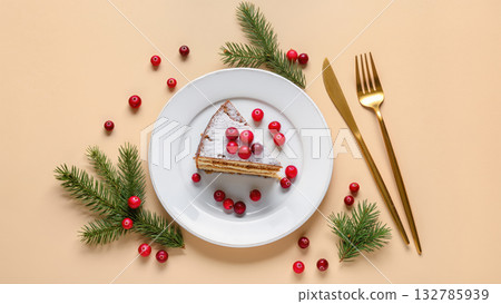 Christmas dessert slice on white plate with cranberries and powdered sugar, decorated with fir branches and golden cutlery on pastel background. Christmas dessert slice on white plate with cranberries and powdered sugar, decorated with fir branches and golden cutlery on pastel background. 132785939