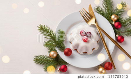 Christmas dessert with white icing and cranberries on a white plate, surrounded by fir branches, baubles and golden cutlery. Festive holiday treat. 132785940