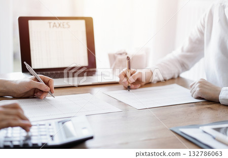 Two accountants using a laptop computer and calculator while counting taxes at wooden desk in office. Teamwork in business audit and finance Two accountants using a laptop computer and calculator while counting taxes at wooden desk in office. Teamwork in business audit and finance 132786063