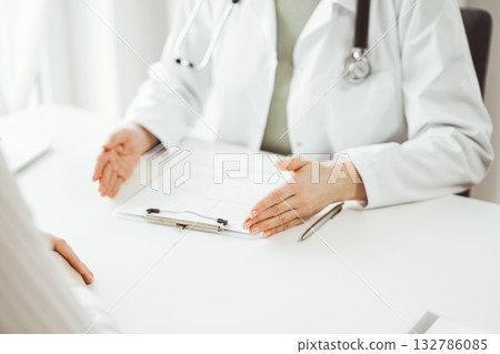 Doctor and patient sitting and discussing something near each other at the white desk in clinic. Female physician is listening filling up a records form. Medicine concept Doctor and patient sitting and discussing something near each other at the white desk in clinic. Female physician is listening filling up a records form. Medicine concept 132786085