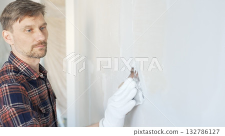 Male construction worker spreading plaster on white wall, wearing protective gloves during home renovation project with putty knife Male construction worker spreading plaster on white wall, wearing protective gloves during home renovation project with putty knife 132786127