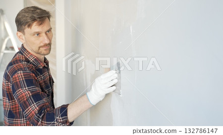 Construction worker man wearing gloves and holding a putty knife is applying plaster on a white wall, performing home renovation and improvement Construction worker man wearing gloves and holding a putty knife is applying plaster on a white wall, performing home renovation and improvement 132786147