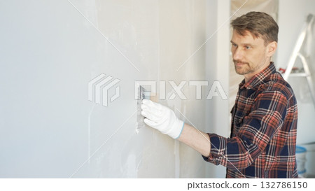 Construction worker man wearing gloves and holding a putty knife is applying plaster on a white wall, performing home renovation and improvement 132786150