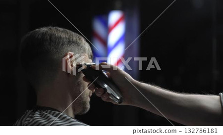 Skilled barber trimming client's hair using electric clipper, vintage barbershop interior with blurred pole, professional grooming service Skilled barber trimming client's hair using electric clipper, vintage barbershop interior with blurred pole, professional grooming service 132786181