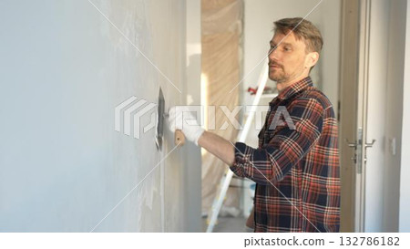 Male construction worker wearing protective gloves smoothing plaster onto wall surface during home renovation using professional trowel tool Male construction worker wearing protective gloves smoothing plaster onto wall surface during home renovation using professional trowel tool 132786182