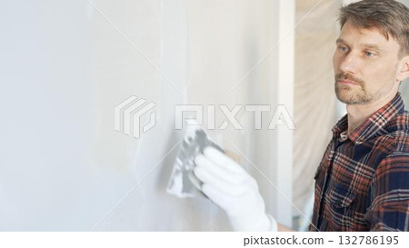 Male construction worker spreading plaster on white wall, wearing protective gloves during home renovation project with putty knife Male construction worker spreading plaster on white wall, wearing protective gloves during home renovation project with putty knife 132786195