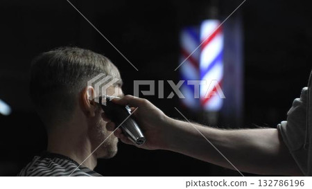 Skilled barber trimming client's hair using electric clipper, vintage barbershop interior with blurred pole, professional grooming service 132786196