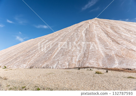 Potassium ore dump. Massive mound potash waste rises deep blue sky, its surface marked by mineral lines erosion patterns. Arid landscape sparse vegetation industrial origin and stark natural contrast 132787593