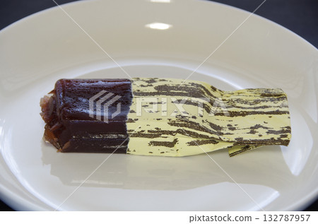 A bite-sized piece of Shimizu Yokan, with its individual wrapper peeled off and half of the contents squeezed out, is placed horizontally on a white plate. 132787957