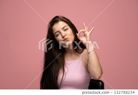 Playful young woman making OK gesture and kissing face on pink background 132788234