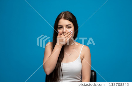 Surprised emotion of young woman covering mouth with hand in white tank top on blue background 132788243