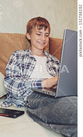 Young man at home smiling with satisfaction, is emotionally working on a laptop. Caucasian guy sitting on sofa and enthusiastically taps on the keyboard.  132788282