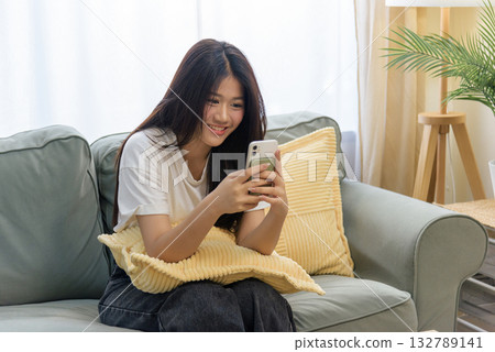 Smiling young Asian woman happy using mobile phone for online communication, person relaxing comfortably on green sofa at home, enjoying cell phone browsing, internet connection, digital lifestyle. Smiling young Asian woman happy using mobile phone for online communication, person relaxing comfortably on green sofa at home, enjoying cell phone browsing, internet connection, digital lifestyle. 132789141