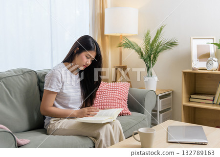 Young woman reading book on sofa, teen person finding rest in comfortable home room, enjoying quiet moment, relaxing with leisure activity, gaining knowledge, long dark hair. Young woman reading book on sofa, teen person finding rest in comfortable home room, enjoying quiet moment, relaxing with leisure activity, gaining knowledge, long dark hair. 132789163