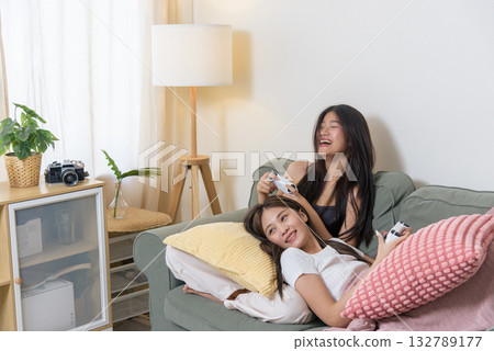 Two young teen friends play exciting videogame at home, laughing heartily together, enjoying immersive console gaming activity, happy teenagers having fun with white controllers on soft green sofa. 132789177