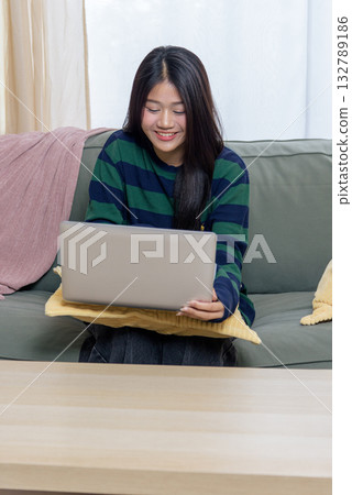 Smiling teen woman happy at home using laptop for work, customer engagement, shop use, embracing cozy online tasks on comfortable green sofa, remote lifestyle. Smiling teen woman happy at home using laptop for work, customer engagement, shop use, embracing cozy online tasks on comfortable green sofa, remote lifestyle. 132789186