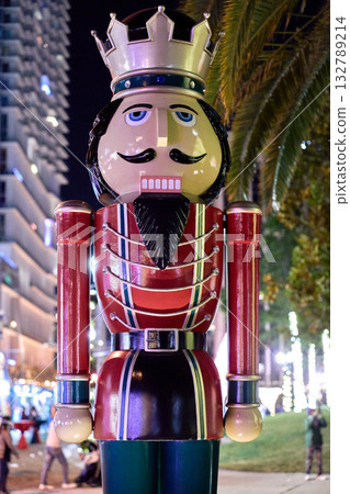 A nutcracker soldier statue with a crown, mustache, and red uniform is lit by bright holiday lights in the city at night. The classic Christmas decoration stands in an outdoor public space with people A nutcracker soldier statue with a crown, mustache, and red uniform is lit by bright holiday lights in the city at night. The classic Christmas decoration stands in an outdoor public space with people 132789214