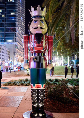 A tall nutcracker soldier statue with a crown stands on a sidewalk in a downtown city at night next to palm trees and holiday lights. People gather in the street behind the festive Christmas 132789215