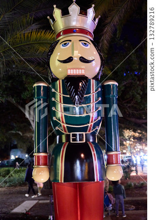 A tall nutcracker soldier statue with a crown, mustache, and bright uniform stands in a park at night under holiday lights. People walk in the background during a festive winter celebration. 132789216