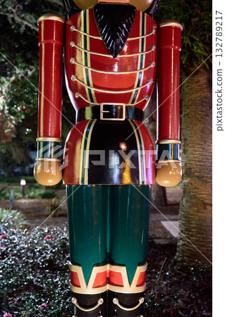 A close-up view shows the shiny red and green uniform of a traditional nutcracker soldier Christmas decoration at night. The belt, buttons, and stripes glow under holiday lights in an outdoor park. A close-up view shows the shiny red and green uniform of a traditional nutcracker soldier Christmas decoration at night. The belt, buttons, and stripes glow under holiday lights in an outdoor park. 132789217