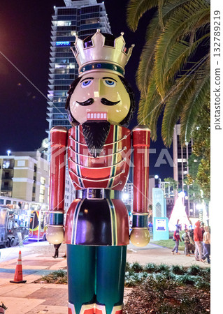 A full-height nutcracker stands on the sidewalk with palms and high-rises. Holiday lights and people create a lively seasonal city scene. A full-height nutcracker stands on the sidewalk with palms and high-rises. Holiday lights and people create a lively seasonal city scene. 132789219