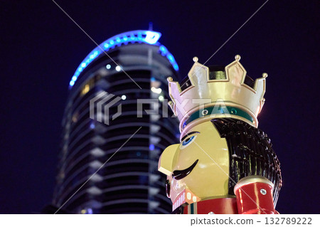 A nutcracker statue in profile stands before a modern blue-lit skyscraper. Urban Christmas decoration glows brightly at night. A nutcracker statue in profile stands before a modern blue-lit skyscraper. Urban Christmas decoration glows brightly at night. 132789222
