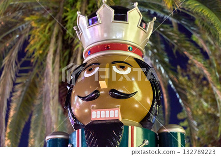 A glowing nutcracker face in Christmas lights is shown close up at night. Festive statue stands by palm trees in a city park. 132789223