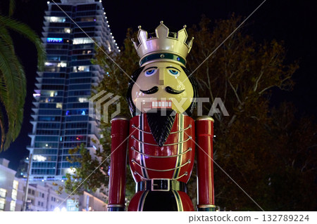 A red nutcracker holiday statue stands under city skyscrapers at night. Festive Christmas decoration adds color to the urban plaza. A red nutcracker holiday statue stands under city skyscrapers at night. Festive Christmas decoration adds color to the urban plaza. 132789224
