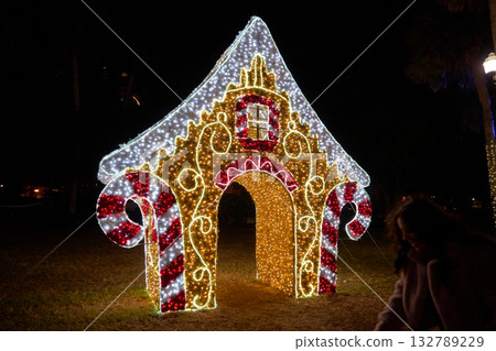 A large gingerbread house arch made of LED lights glows at night. Festive candy cane columns and bright decoration welcome families in a winter park. A large gingerbread house arch made of LED lights glows at night. Festive candy cane columns and bright decoration welcome families in a winter park. 132789229