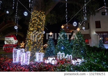 Shimmering present boxes glow beside star-topped trees and a wrapped oak trunk. The park shines with LED lights and cheerful holiday color. 132789251