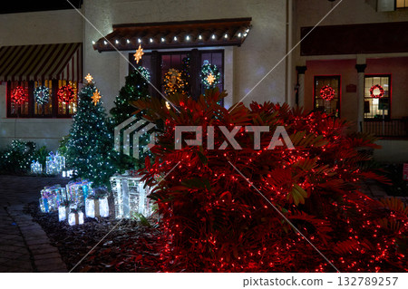 A home exterior shines with wreaths, red-lit bushes, and glowing evergreens at night. Festive holiday decorations line the sidewalk with bright gift boxes. A home exterior shines with wreaths, red-lit bushes, and glowing evergreens at night. Festive holiday decorations line the sidewalk with bright gift boxes. 132789257