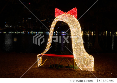 Illuminated arch with a red bow and bench glows beside the city lake at night. Festive holiday decoration with bright LED lights and golden sparkle. 132789260