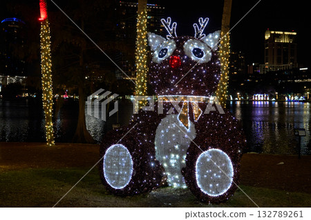 Large reindeer figure made of LED lights glows beside a city lake at night. Festive holiday decoration with red nose, antlers, and sparkling details. Large reindeer figure made of LED lights glows beside a city lake at night. Festive holiday decoration with red nose, antlers, and sparkling details. 132789261