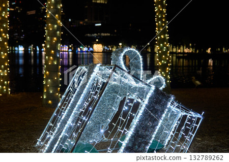 Sparkling present box with a glowing bow stands by a lake at night. White and green LED lights create a festive holiday display. 132789262