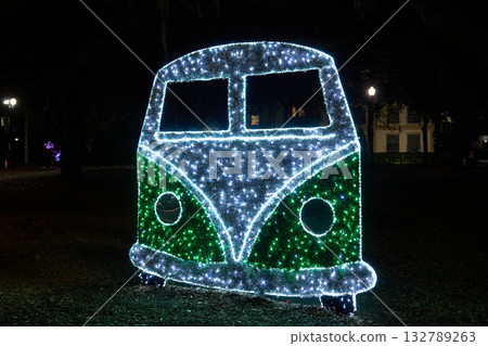 Large illuminated camper van silhouette glows with green and white LED lights in a park. Festive holiday decoration with vintage bus style and sparkling details. Large illuminated camper van silhouette glows with green and white LED lights in a park. Festive holiday decoration with vintage bus style and sparkling details. 132789263