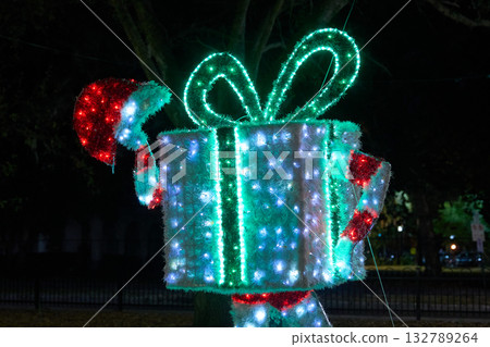 Large illuminated gift box with ribbon, bow, and Santa hat stands glowing in a park at night. Festive holiday display with bright LED lights and colorful accents. Large illuminated gift box with ribbon, bow, and Santa hat stands glowing in a park at night. Festive holiday display with bright LED lights and colorful accents. 132789264