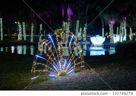 A fan of warm and cool LED rods glows near a pond with lit trees and fountains. The outdoor installation adds festive decoration to the city park. A fan of warm and cool LED rods glows near a pond with lit trees and fountains. The outdoor installation adds festive decoration to the city park. 132789279