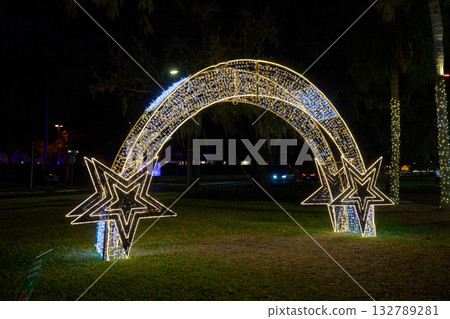 A curved arch with star ends glows with LED lights on a grassy park at night. The holiday installation forms a bright entrance for a festive celebration. A curved arch with star ends glows with LED lights on a grassy park at night. The holiday installation forms a bright entrance for a festive celebration. 132789281