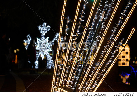 Tall golden and white LED rods glow beside a pathway at night. A bright snowflake display shines in the background, creating a festive holiday atmosphere. 132789288