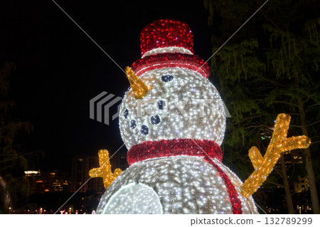 A bright LED snowman with a red hat and scarf glows at night. Holiday decoration brings cheerful winter spirit to the city. A bright LED snowman with a red hat and scarf glows at night. Holiday decoration brings cheerful winter spirit to the city. 132789299