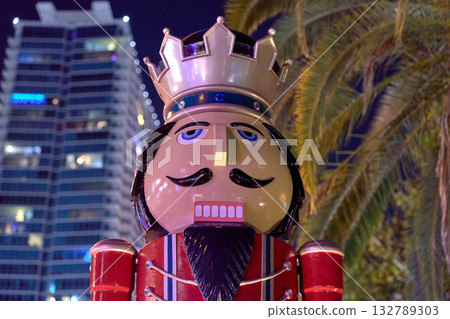 A bright nutcracker figure stands near palm trees and a high-rise building at night. Festive Christmas decoration adds cheerful holiday spirit to the city. A bright nutcracker figure stands near palm trees and a high-rise building at night. Festive Christmas decoration adds cheerful holiday spirit to the city. 132789303