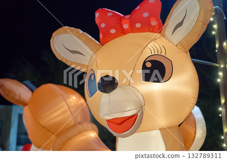 A cheerful inflatable doe wears a red polka-dot bow and smiles toward the camera. The glowing reindeer stands outdoors beneath twinkling lights. 132789311