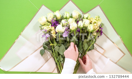 Hands gently adjust floral stems within a large bouquet of white, purple, and yellow blooms. The bouquet is positioned on wrapping paper against a green background. Hands gently adjust floral stems within a large bouquet of white, purple, and yellow blooms. The bouquet is positioned on wrapping paper against a green background. 132789444