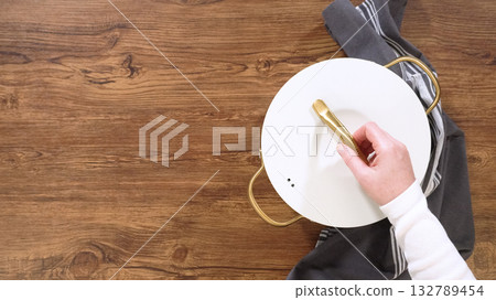 A person places a white lid on a ceramic pot resting on a striped towel. The pot has gold handles and sits on a wooden surface. 132789454