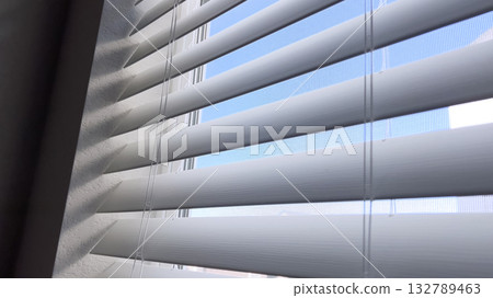 Natural light shines through partially open white window blinds. The view includes a soft blue sky and neutral-colored walls. 132789463