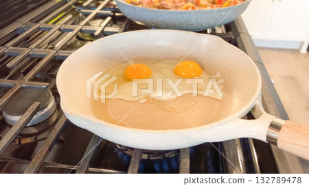 Two cracked eggs fry in a light-colored ceramic skillet over a gas flame. A pan of breakfast hash sits on the burner behind. Two cracked eggs fry in a light-colored ceramic skillet over a gas flame. A pan of breakfast hash sits on the burner behind. 132789478