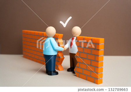 Two businessmen shake hands through a gap in a brick wall. To find common ground and begin negotiations for a compromise. Teamwork, partnership, mediation, or business deal negotiations concepts. Two businessmen shake hands through a gap in a brick wall. To find common ground and begin negotiations for a compromise. Teamwork, partnership, mediation, or business deal negotiations concepts. 132789663